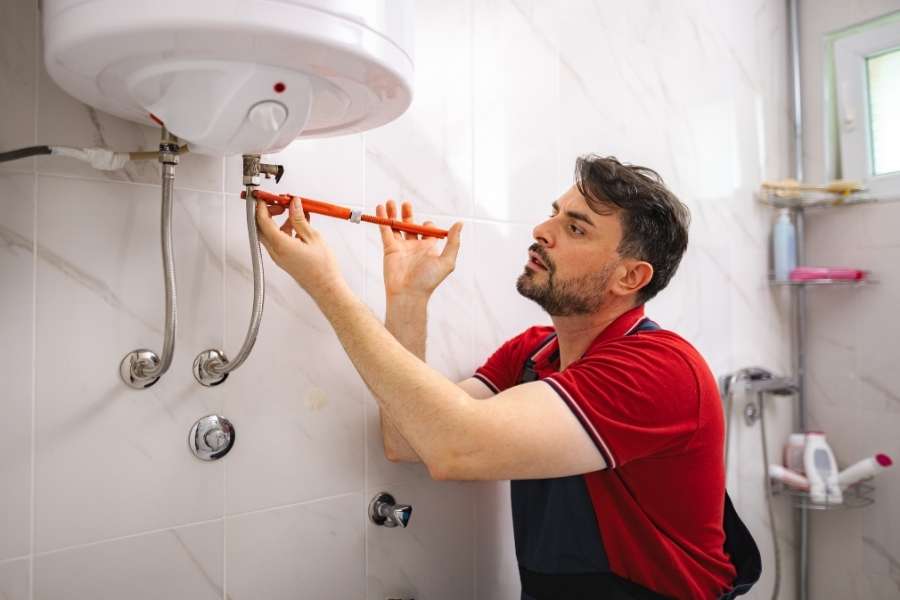 Un plombier concentré, portant un t-shirt rouge et une combinaison, serre un raccord sur la tuyauterie d'un chauffe-eau thermodynamique mural dans une salle de bain carrelée.